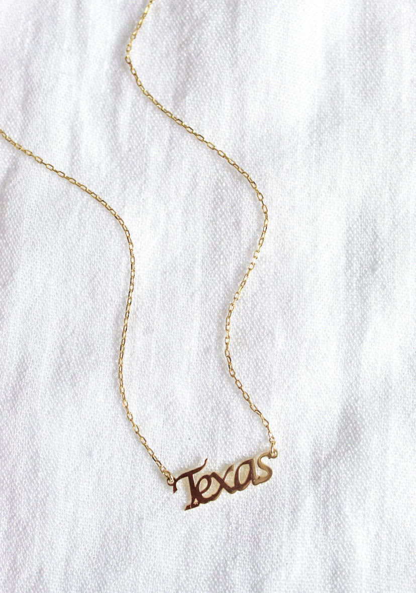 The Kinsey Designs Archives State of Mind Necklace features a gold filled pendant hand stamped with "Texas," displayed on a white textured fabric background.