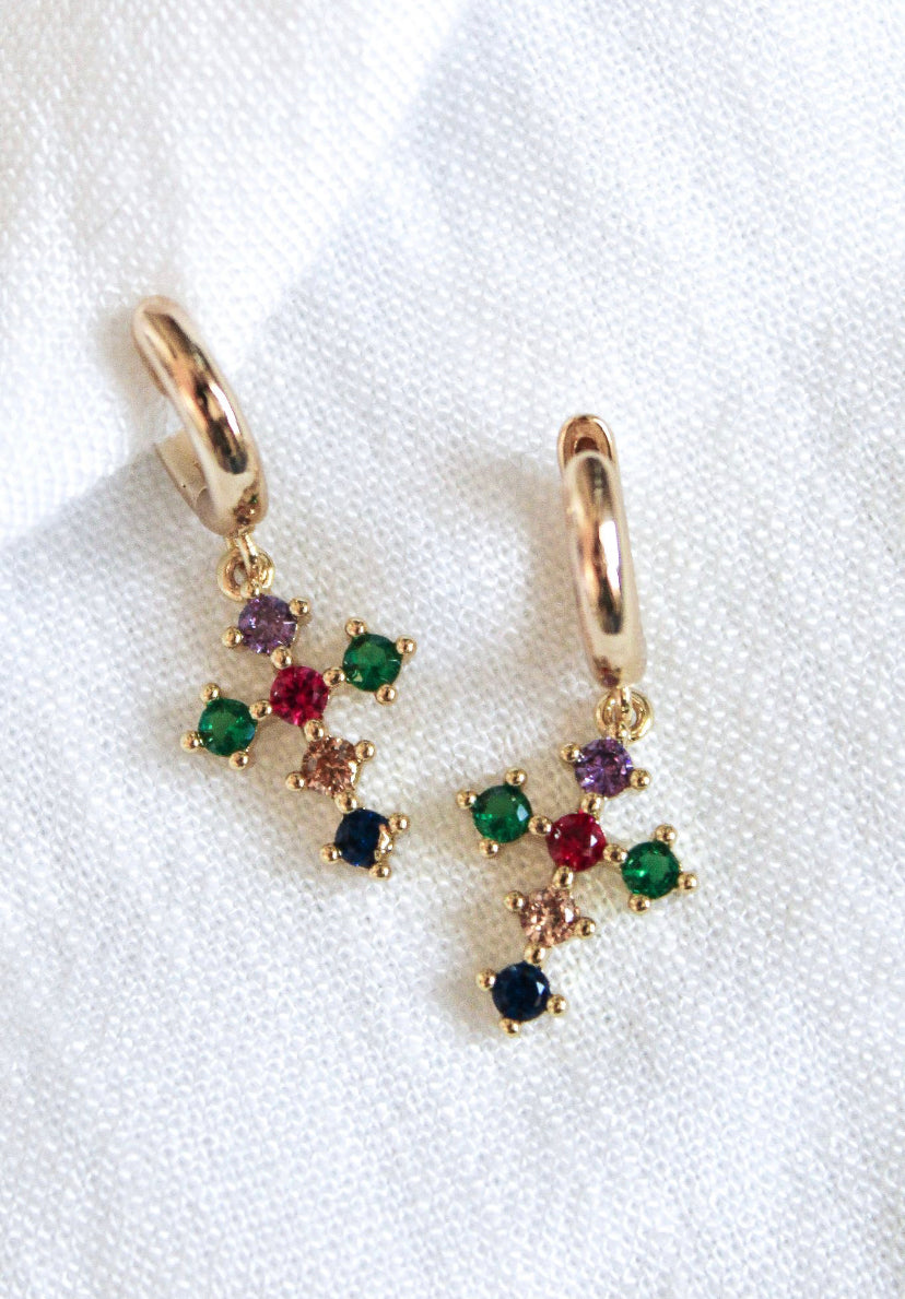 The Tori Huggie - Multi by Kinsey Designs Archives features gold filled hoops with dangling multicolored cross charms, displayed on a white textured fabric background.