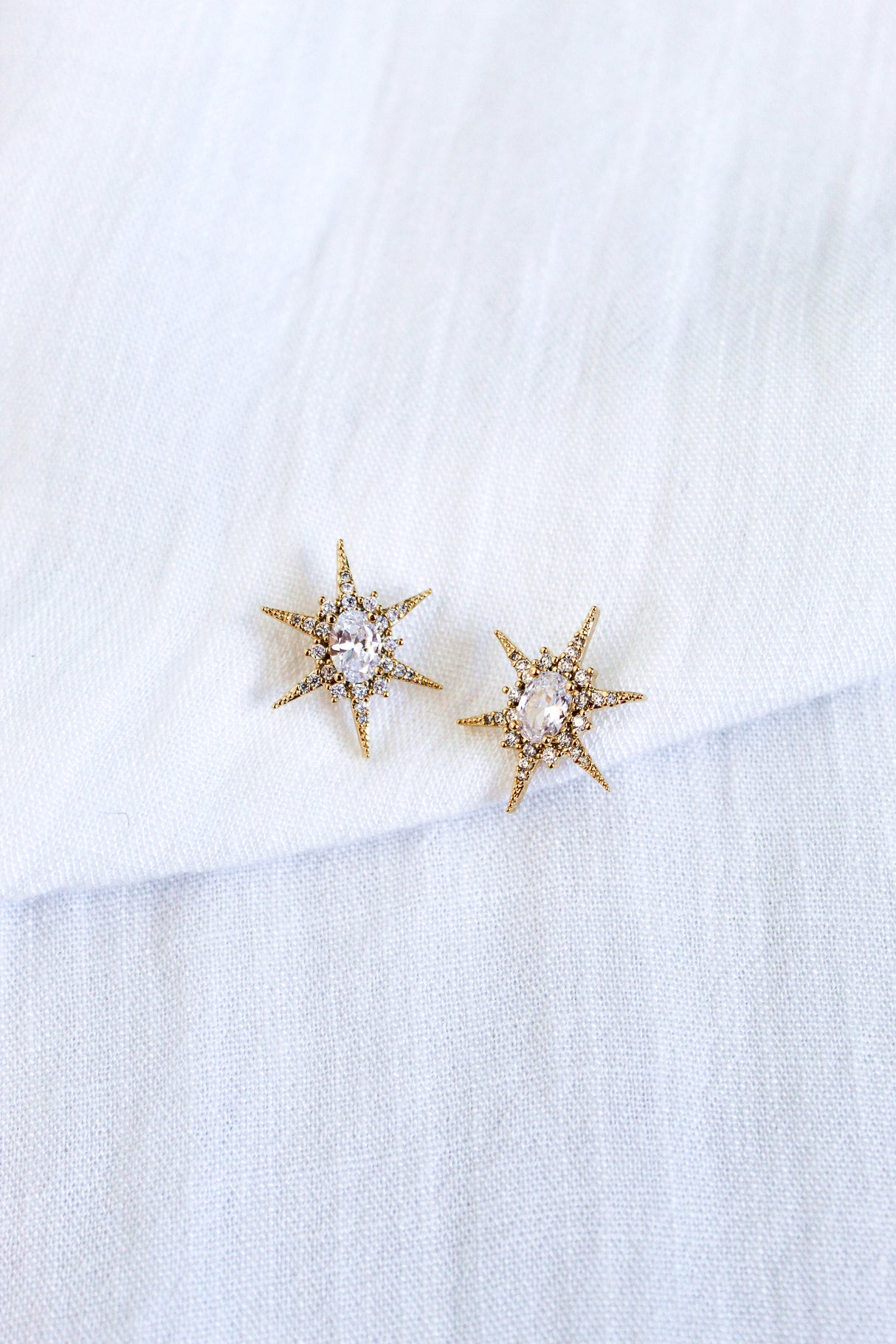 The Faye Stud by Kinsey Designs features gold star-shaped earrings with clear cubic zirconia gemstones in gold-filled metal, showcased on a white textured fabric background.