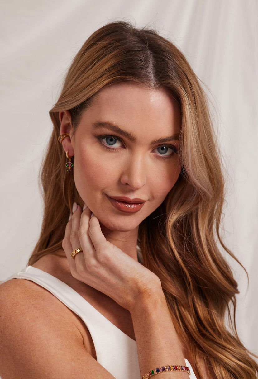 A woman with long wavy hair wears Kinsey Designs Archives' Tori Huggie - Multi gold filled earrings, posing with her hand near her face against a light backdrop.