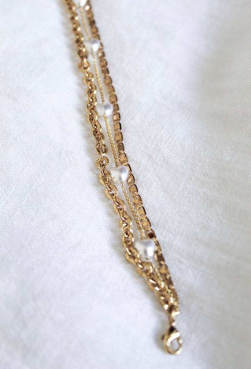 The Beyah Layer Bracelet by Kinsey Designs Archives is a gold-plated brass piece with two rows and small pearls, shown on a light textured fabric background.