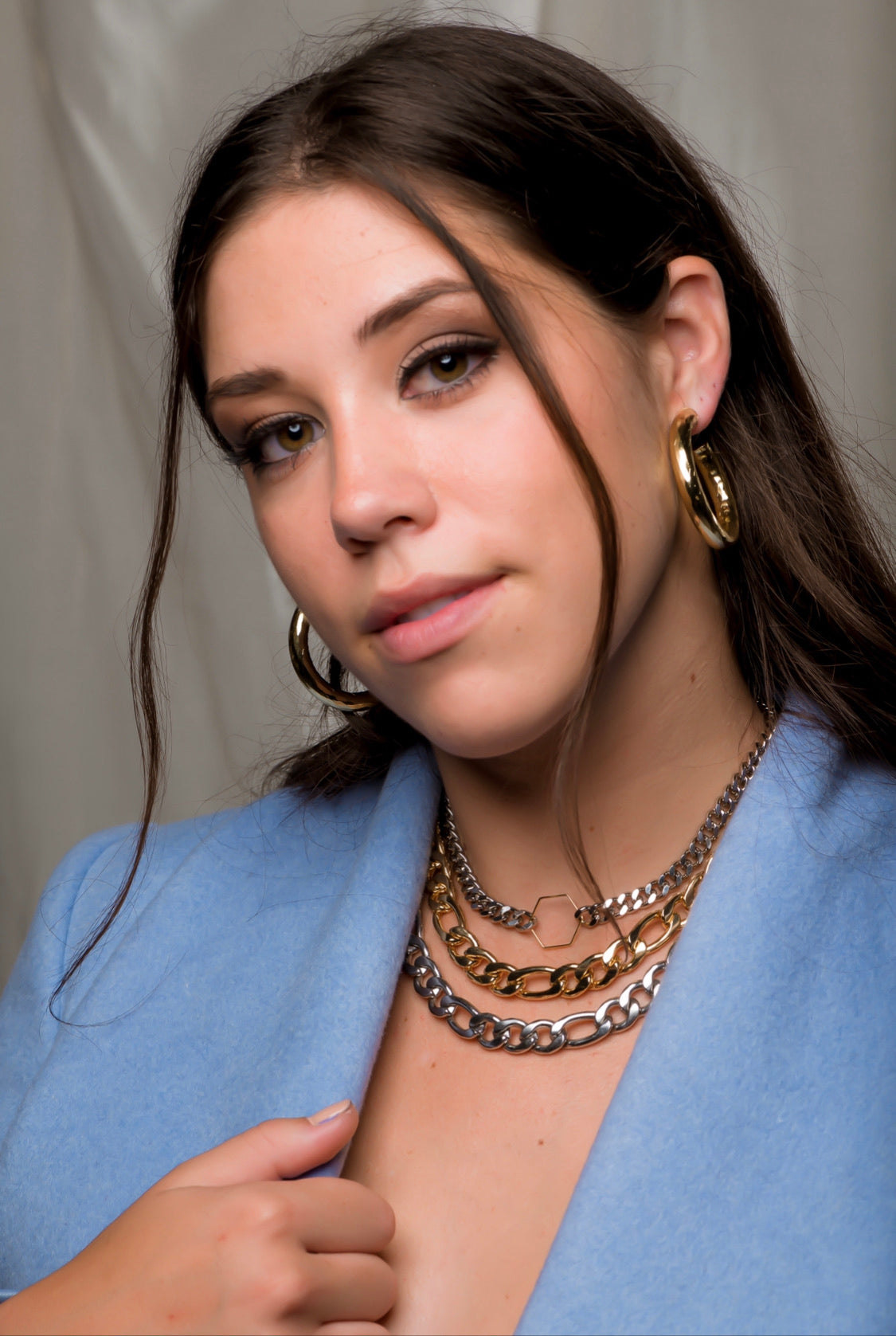 A woman with long brown hair wears Kinsey Designs' Jasmine Hoops, layered necklaces, and a blue jacket, showcasing statement jewelry indoors.