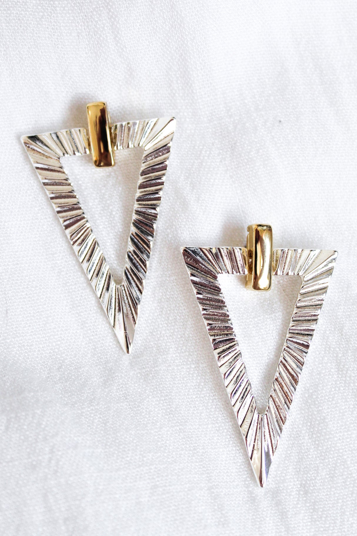 The Colin Earring by Kinsey Designs features silver triangular gold-filled earrings with textured lines and gold-colored top accents, displayed on white fabric. Perfect for a chic look—shop the sample sale now!.