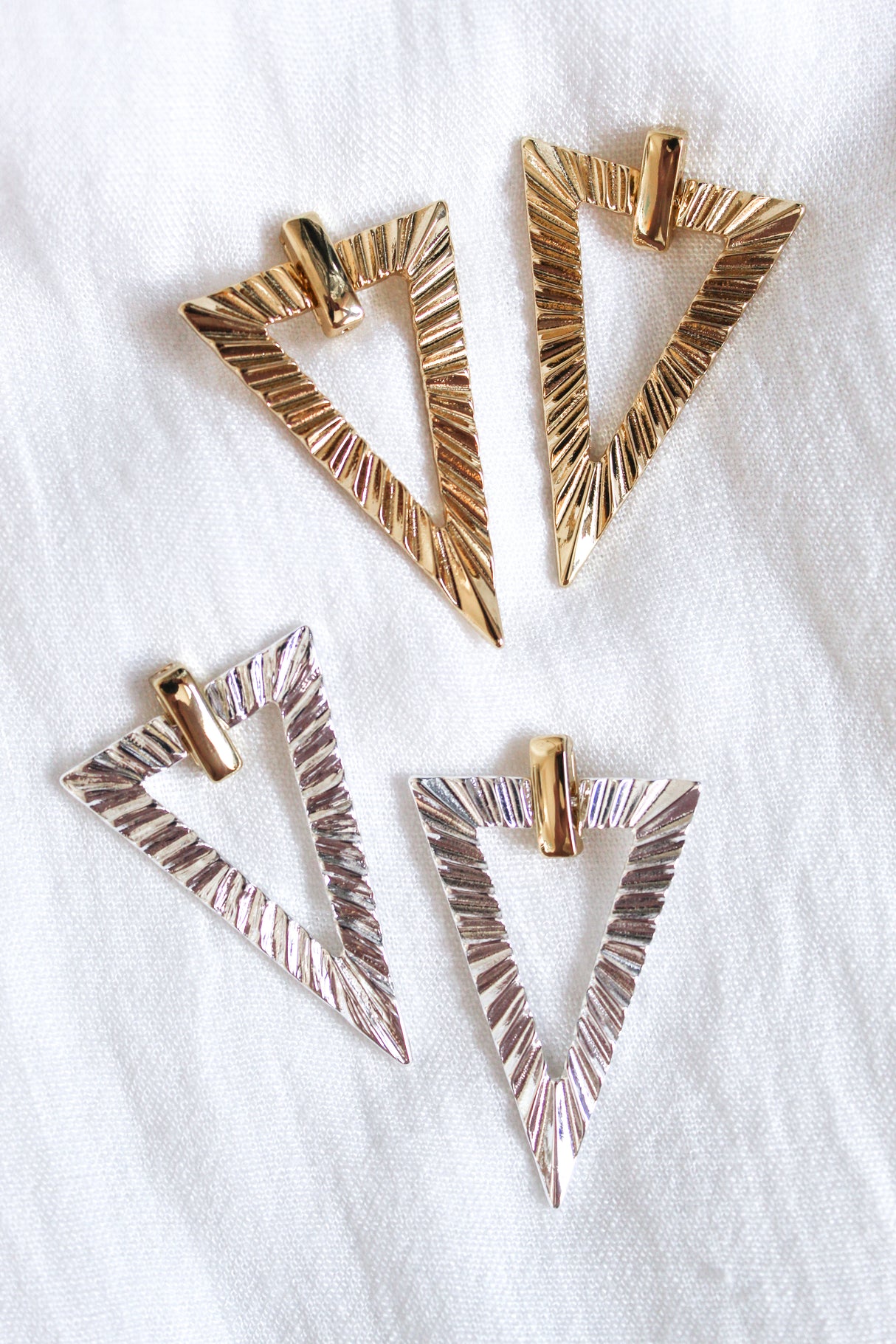 Four textured, triangular metal Colin Earrings by Kinsey Designs—two gold, two silver—styled on a white fabric background. Ideal for collecting or gifting at our exclusive sample sale.
