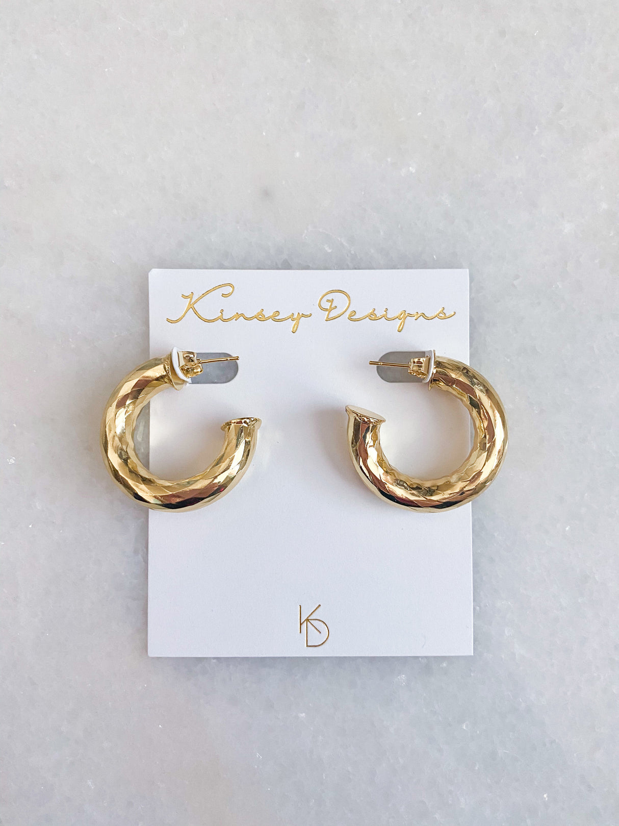 The Jasmine Hoops by Kinsey Designs feature a hammered gold finish, displayed on a white card atop a marble surface—stylish statement earrings for any occasion.