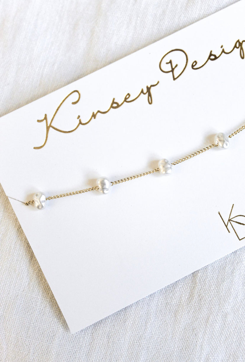 The Adalee gold chain bracelet by Kinsey Designs Archives features small white pearls and is displayed on a branded card. Pair it with a gold plated necklace for a coordinated look.
