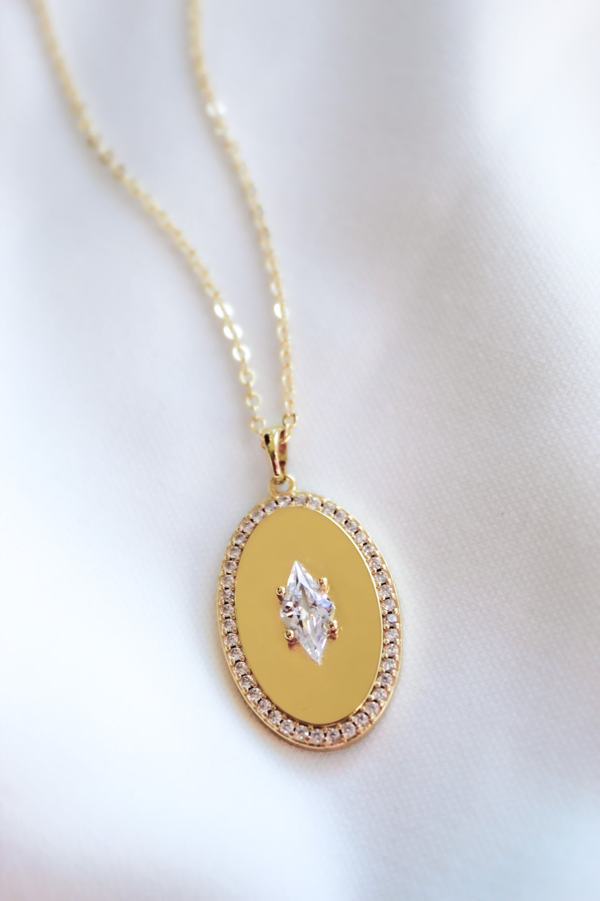 The Fique Necklace by Kinsey Designs features a gold-plated 32-inch chain with a central cubic zirconia pendant encircled by sparkling stones, beautifully displayed on white fabric—perfect for elegant layering.