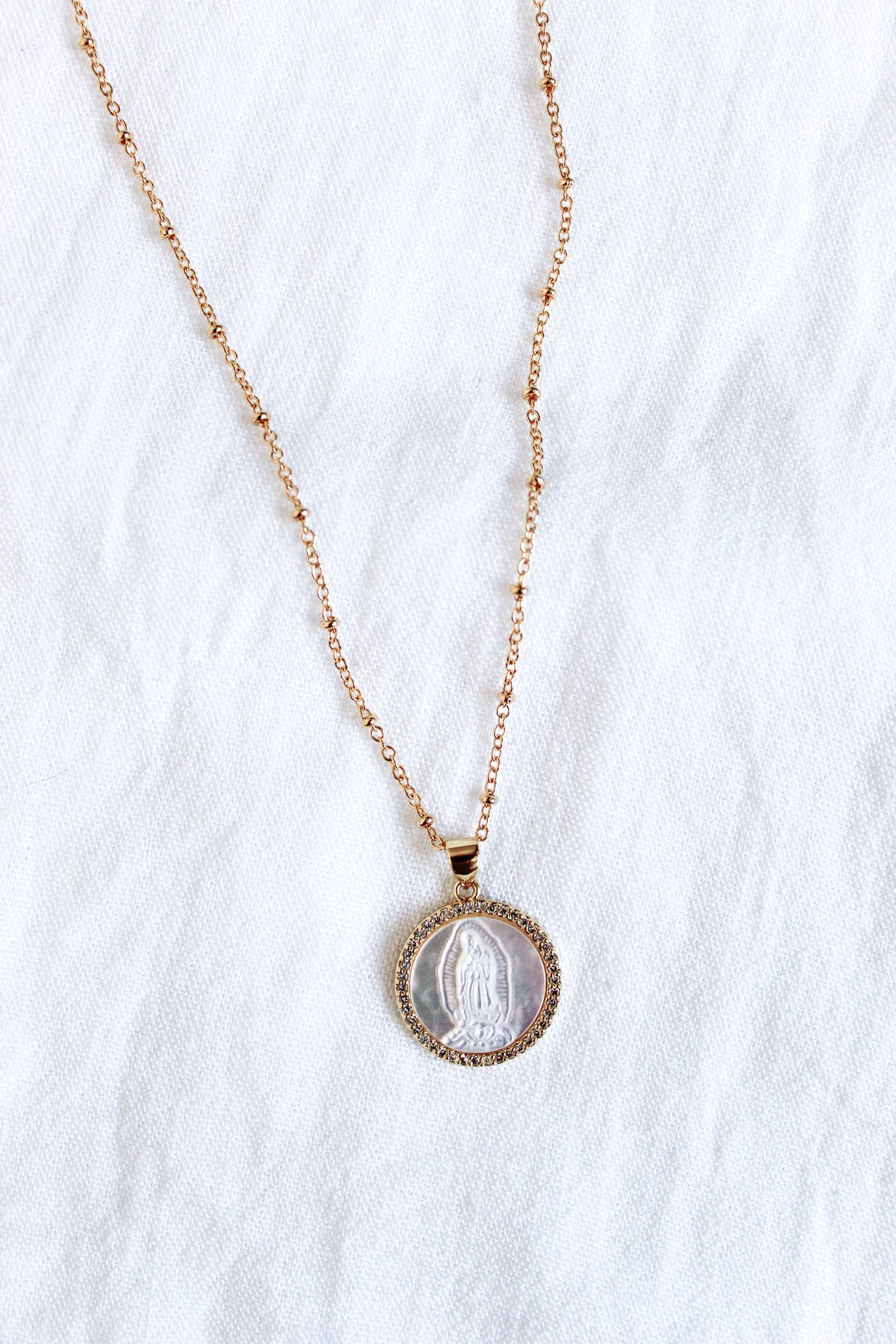 The Alivia necklace by Kinsey Designs Archives features a 14k gold plated chain and an oval pendant with an engraved figure, displayed on a white fabric background.