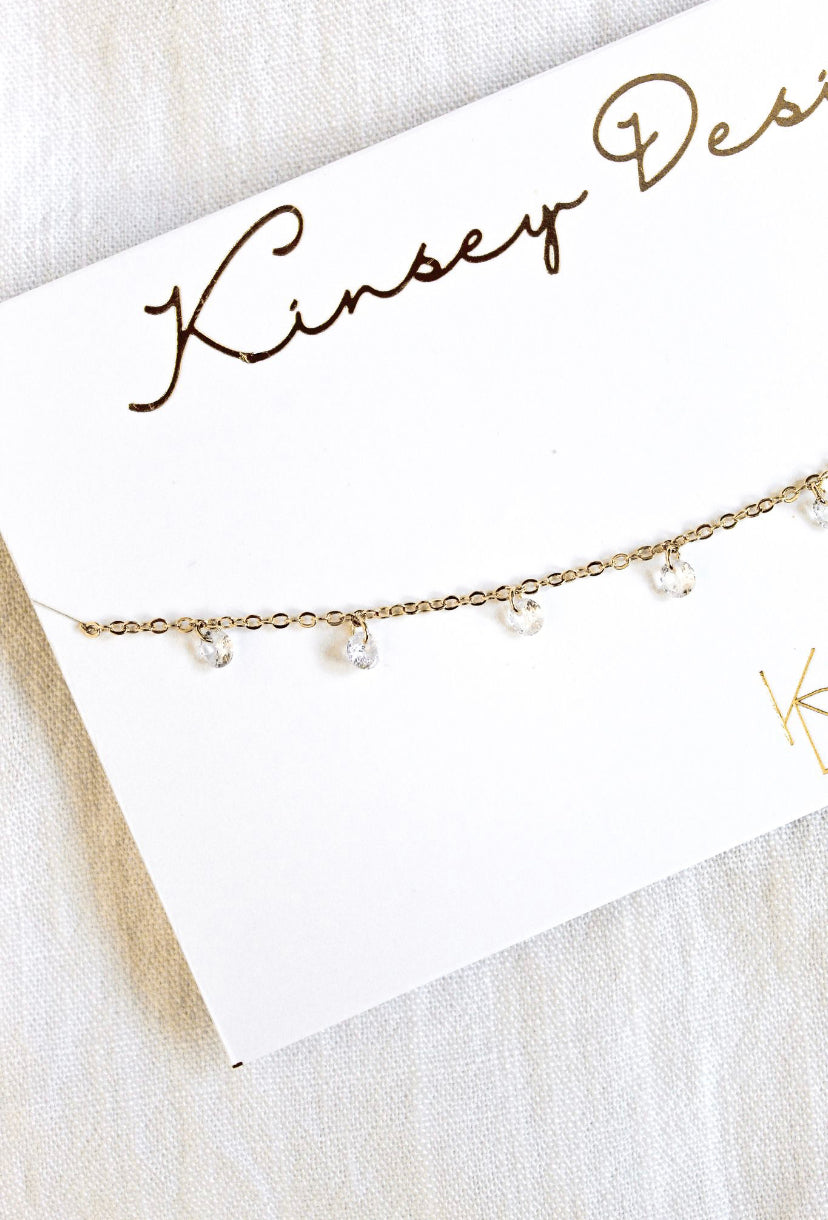 The Clare Choker by Kinsey Designs Archives features a gold plated 14-inch chain with small clear cubic zirconia pendants, displayed on a branded card.
