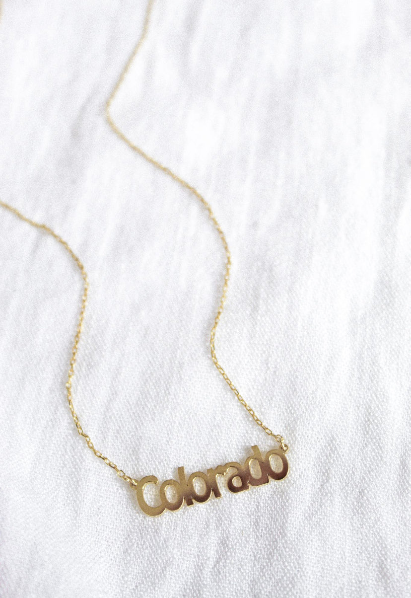 The Kinsey Designs Archives State of Mind Necklace features a "Colorado" pendant, shown on white fabric. Celebrate your hometown pride with this timeless piece from our State of Mind collection.