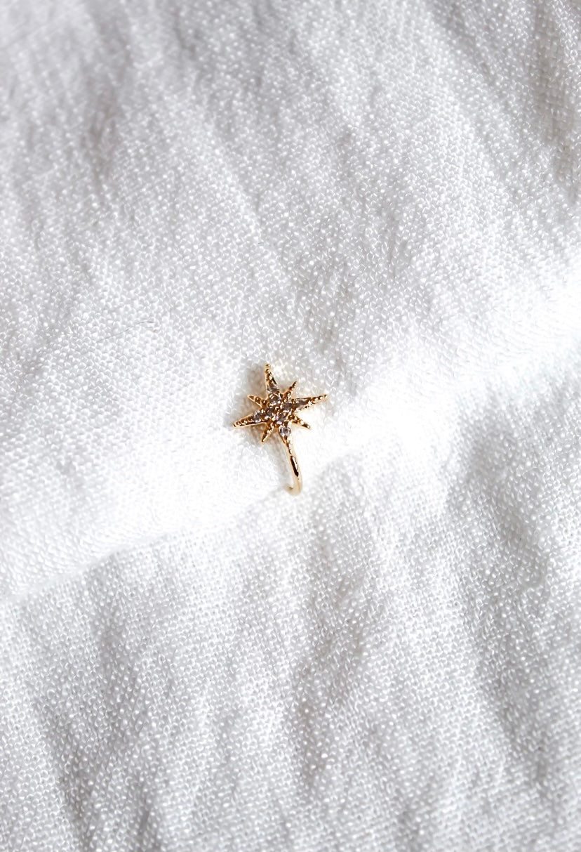 The Burst Ear Cuff by Kinsey Designs Archives is a gold plated, star-shaped ear cuff adorned with sparkling cubic zirconia, shown on white textured fabric.