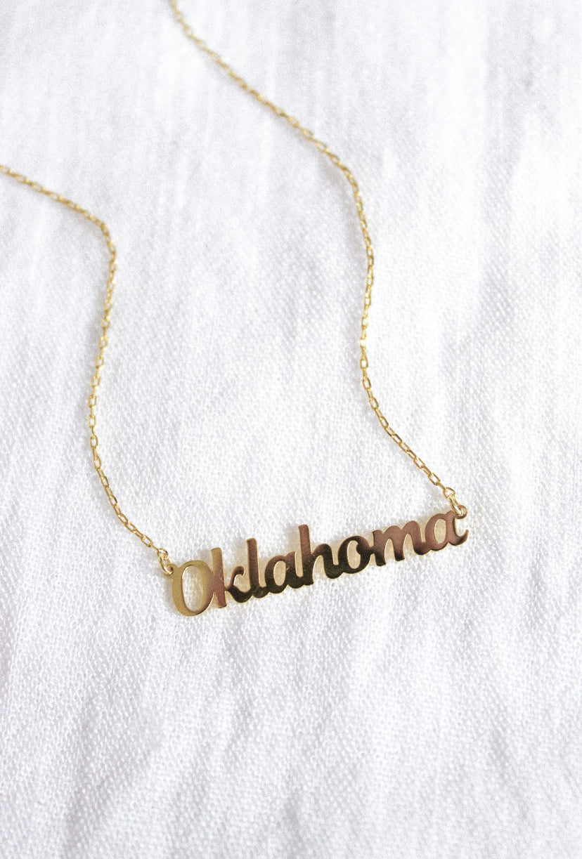Kinsey Designs Archives State of Mind Necklace in gold fill, featuring "Oklahoma" in cursive on a delicate chain, shown on white fabric.