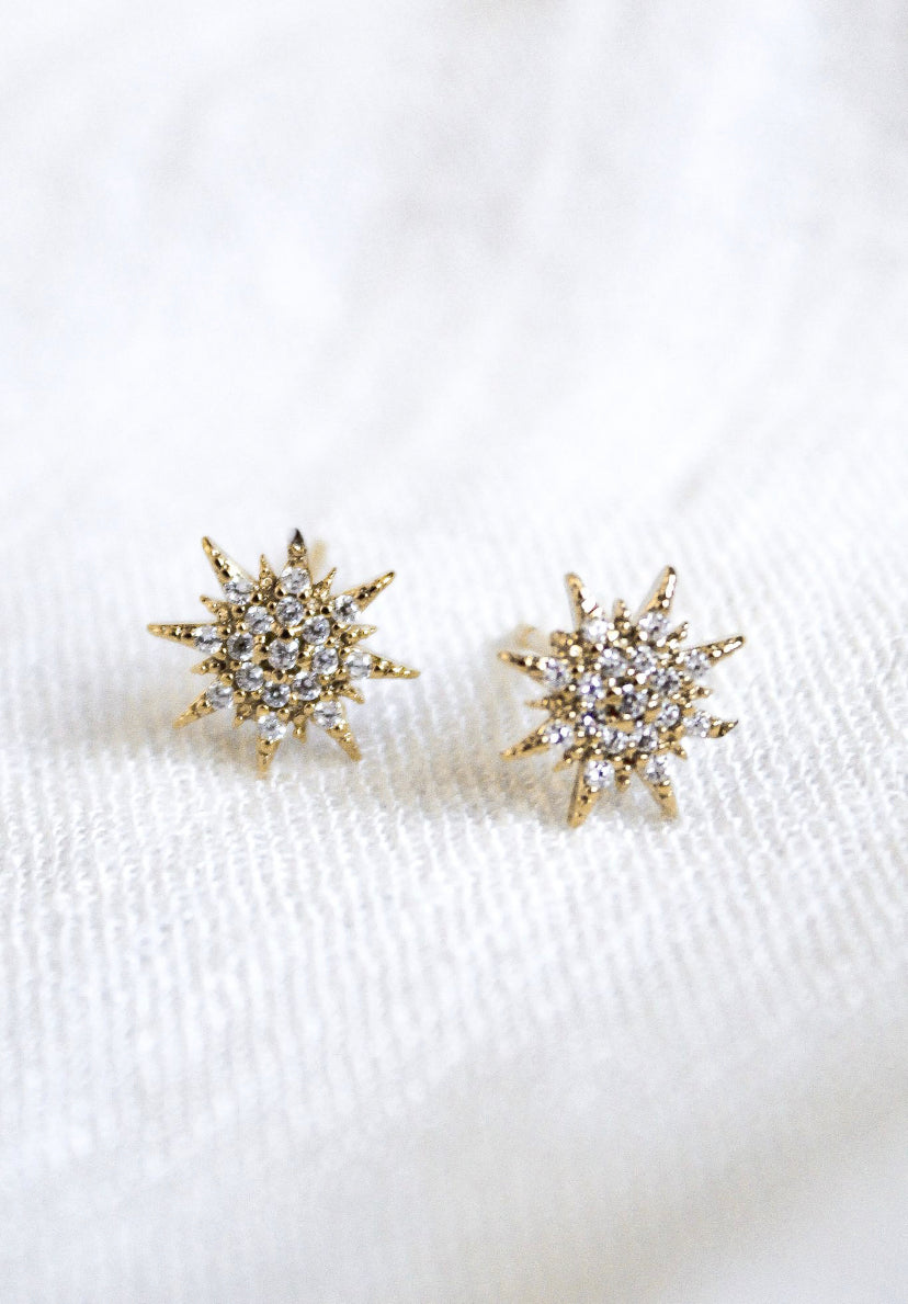 Ace by Kinsey Designs Archives: 16K gold plated starburst stud earrings with small clear stones, shown on a soft white textured surface—perfect for effortless everyday style.