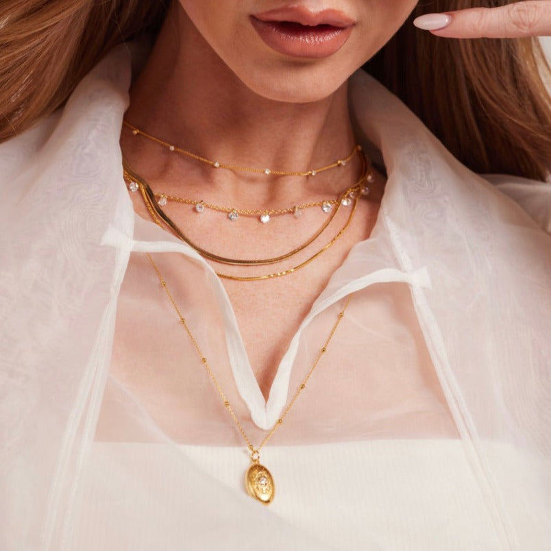 A woman wears the Aria necklace by Kinsey Designs Archives, featuring layered gold plating, a locket, and a 14-inch chain with lobster clasp, styled with a white sheer blouse and nude lipstick.