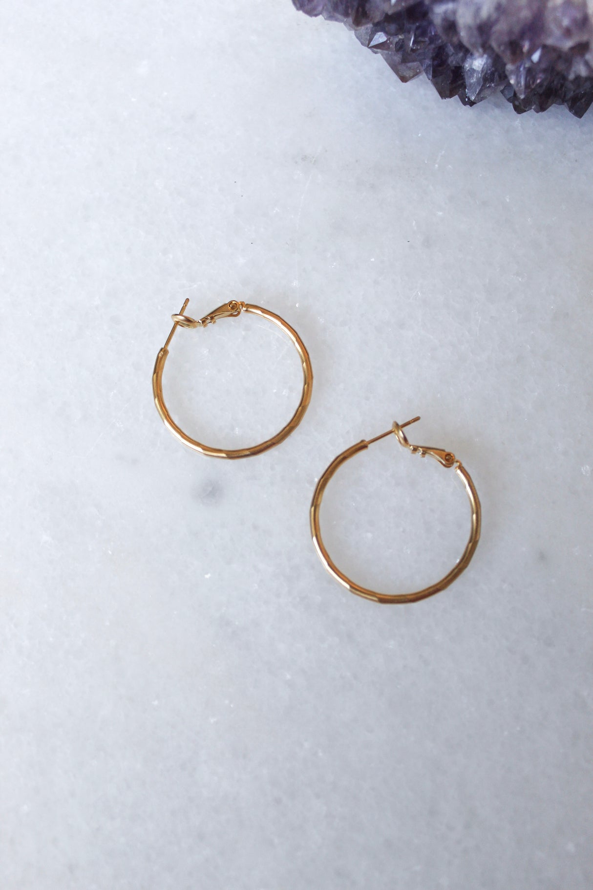 Two small Lexie 18k gold plated hoop earrings by Kinsey Designs Archives rest on a white marble surface, with a purple crystal in the corner.