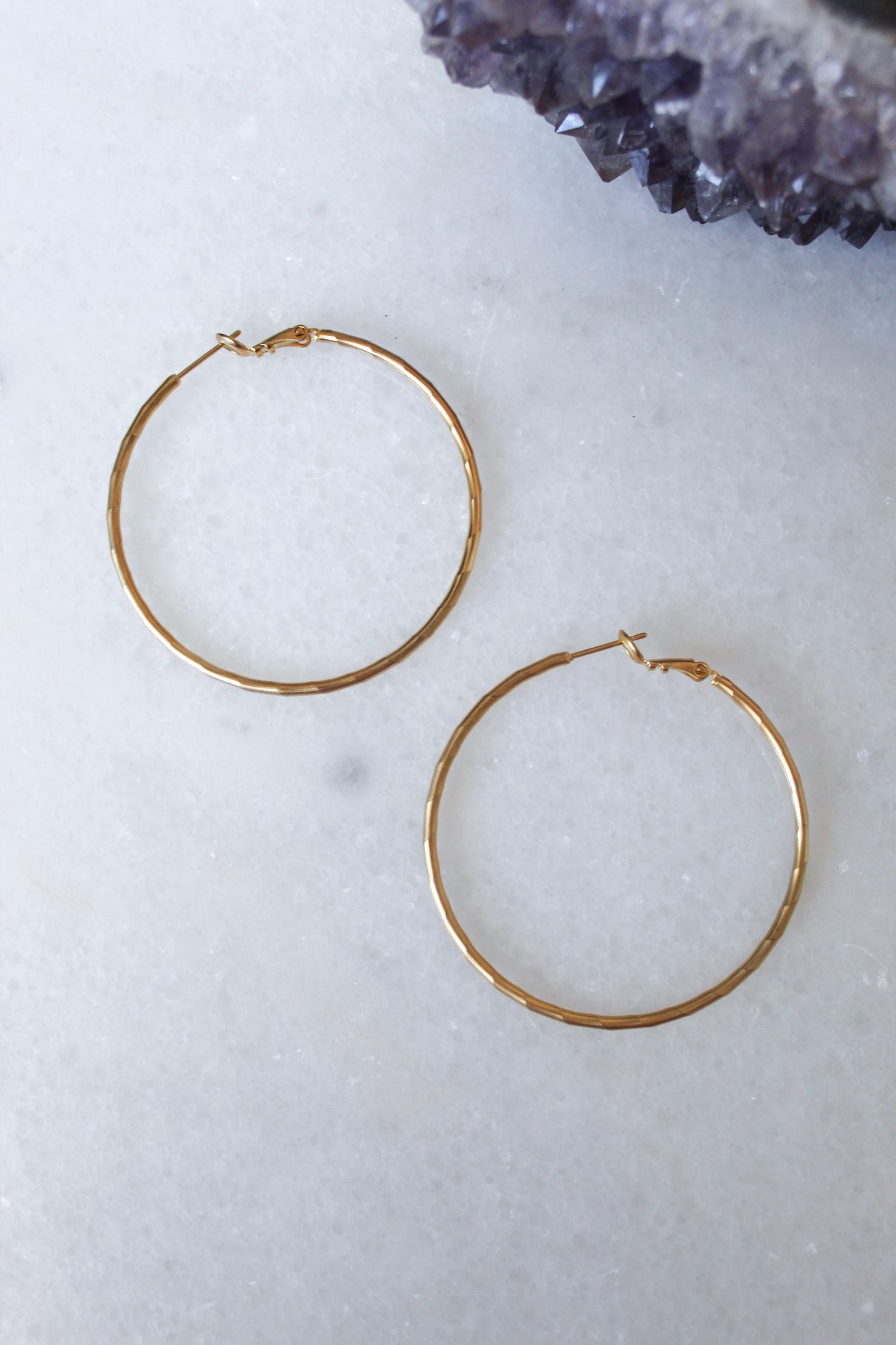 The Lexie earrings by Kinsey Designs Archives—one large 2” and one small 1”, both 18k gold plated hoops—are displayed on a white marble surface with purple crystals in the top right corner.