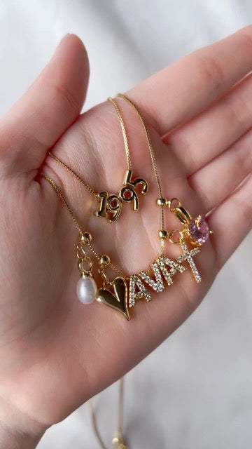 Close-up of a hand holding three gold-filled necklaces with pendants: "1995," a Vintage Heart Drop Charm - Kinsey Charm Bar with "Avani," and a charm bar chain with a pink stone.