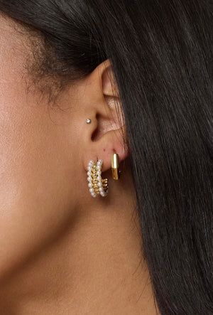 Close-up of an ear with three earrings: gold hoop earrings, shimmering pearl hoops like the Kinsey Designs Archives Elsie Hoop, and a small silver stud.