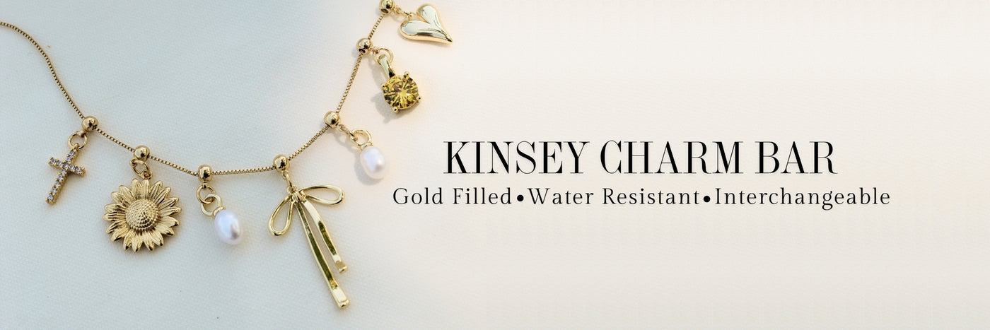 Gold charm bracelet with various charms: cross, sunflower, bow, pearls, heart, and crown. Text reads Kinsey Charm Bar.