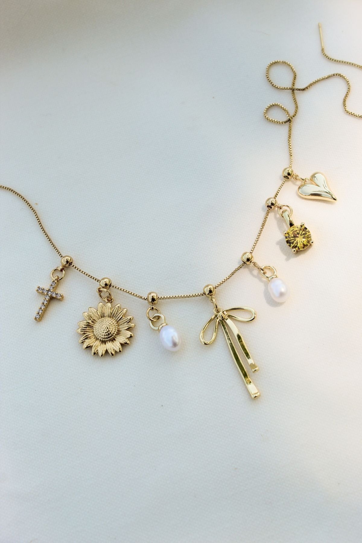 Gold charm necklace with various pendants including a cross, sunflower, pearls, a bow, and a heart.