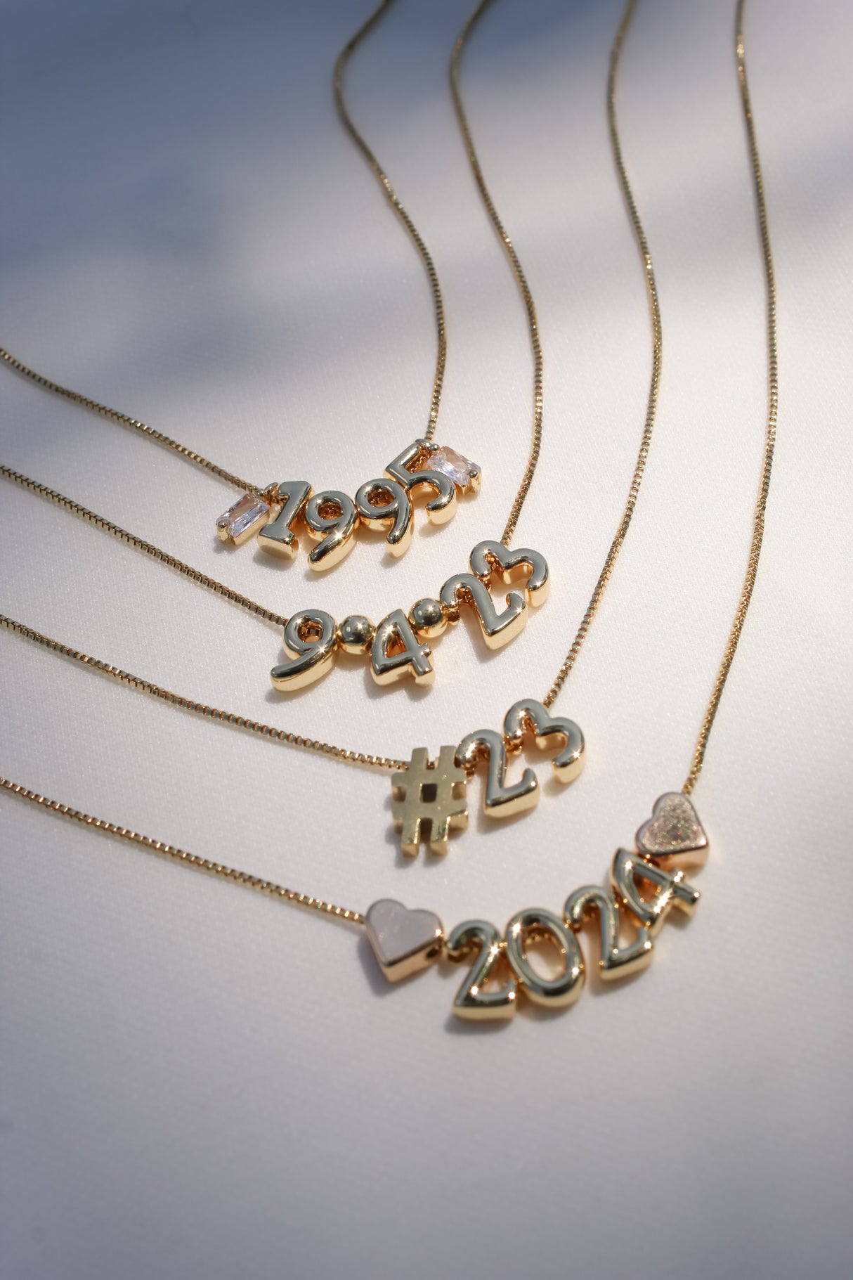 Gold necklaces with pendants displaying the years "1995," "1998," "1994," "#23," and year "2024" on a white background, featuring designs from the Number Symbol Spacer Slide collection and Kinsey Charm Bar.