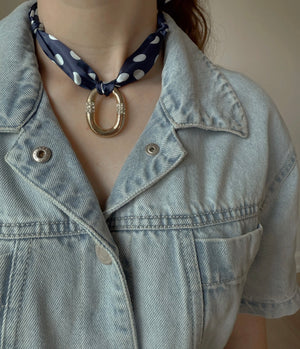 A person wearing a light denim jacket and a navy polka dot adjustable scarf pairs it with the Bellagio Scarf Necklace by Kinsey Designs, featuring a vintage silver horseshoe-shaped pendant.