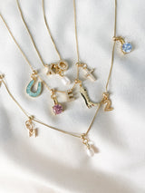 The NEW! Charm Bar Necklace Dainty Cable Chain - Kinsey Charm Bar, featuring various gold-filled necklaces with charms like letters, a horseshoe, a high heel, and gemstones, is elegantly displayed on a white cloth.