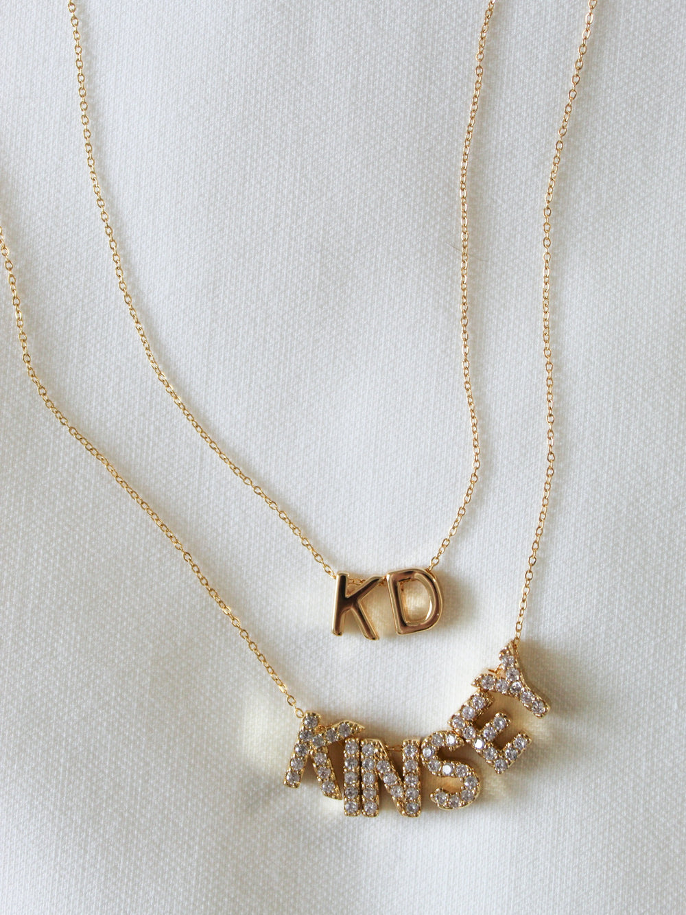 Two gold-filled necklaces from the NEW! Charm Bar Necklace Dainty Cable Chain - Kinsey Charm Bar collection: one with "KD" and the other spelling out "KINSEY" in sparkling letters, both set against a white fabric background. These stunning pieces add a touch of elegance to any outfit.