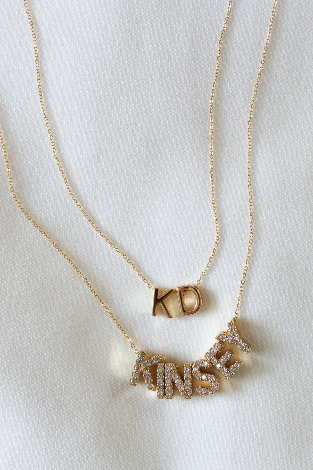 Two gold-filled necklaces from the NEW! Charm Bar Necklace Dainty Cable Chain - Kinsey Charm Bar collection: one with "KD" and the other spelling out "KINSEY" in sparkling letters, both set against a white fabric background. These stunning pieces add a touch of elegance to any outfit.