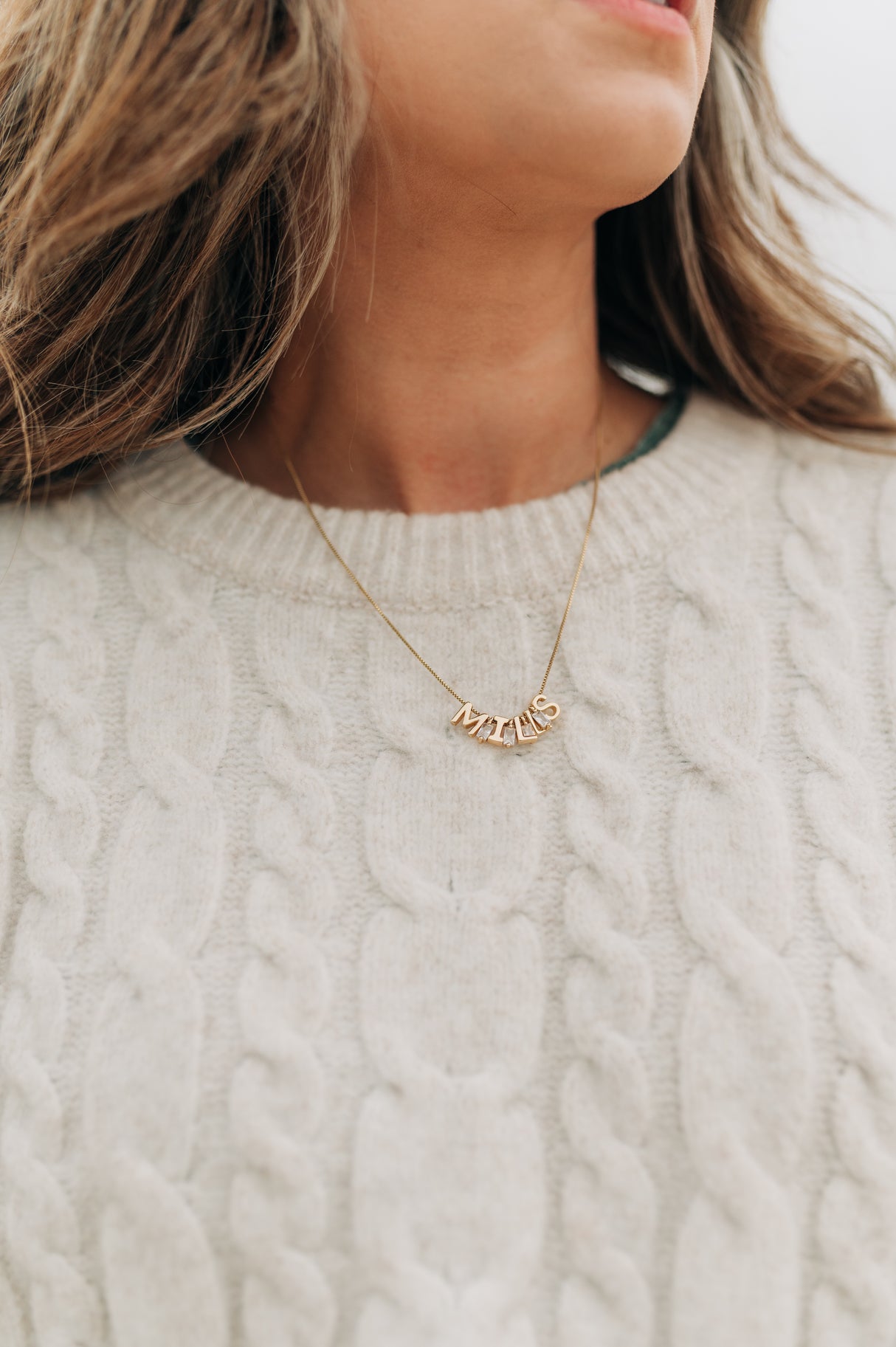 Close-up of a person wearing a beige cable-knit sweater and an adjustable gold-filled Kinsey Charm Bar Necklace Box Chain with the word "MAMA" written in block letters.