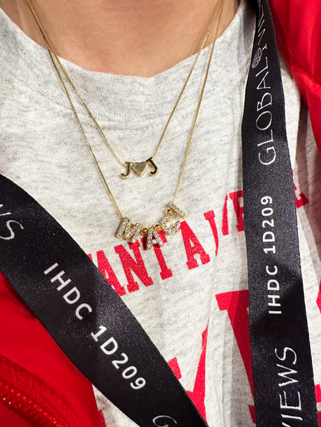 Close-up of layered gold necklaces with letter pendants, a Kinsey Designs Round Spacer Slide accent, and a black lanyard around the neck.