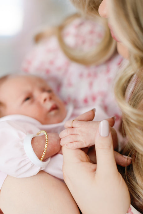 A woman gently holds a newborn’s hand, both adorned with Kinsey Designs’ Fallon Baby Cuff—gold filled and nickel free—in this close-up.