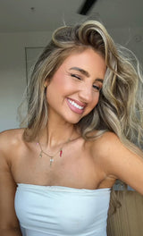 A woman with wavy blonde hair smiles and winks, wearing a white top, gold necklaces, and the Spicy Grip Charm bracelet by Kinsey Designs Archives.