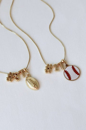 Two gold necklaces from Kinsey Designs' Charm Bar Necklace collection feature number charms, the Football Grip Charm, and a baseball pendant, all displayed on a white background.