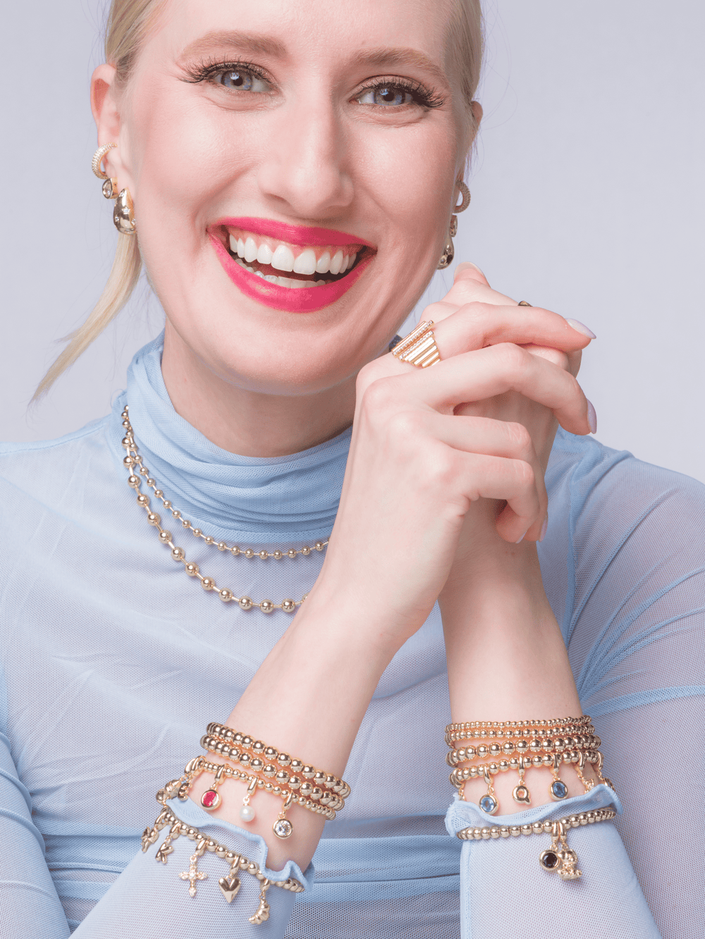 A smiling woman in a sheer blue top wears gold jewelry and colorful Kinsey Designs clip charms, including the Eiffel Tower Clip Charm featuring a secure hinged clasp.