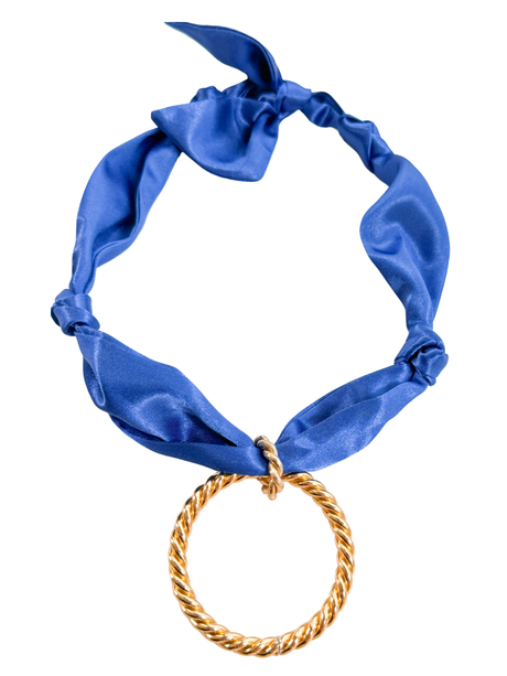 The Arezzo Scarf Necklace by Kinsey Designs is a blue satin necklace featuring a large, twisted gold ring at the center for a chic, adjustable look.