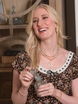 Smiling woman with long blonde hair holds a decorative glass perfume bottle, her outfit accented by the Aurelium Cross Gemstone Necklace from Kinsey Designs, in a cozy room.