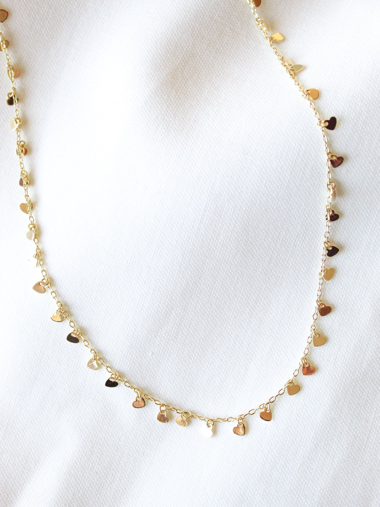 The Avery Necklace by Kinsey Designs Archives features gold-filled small heart-shaped charms on a white textured fabric background. This 14-inch necklace includes an extender for an adjustable, perfect fit.