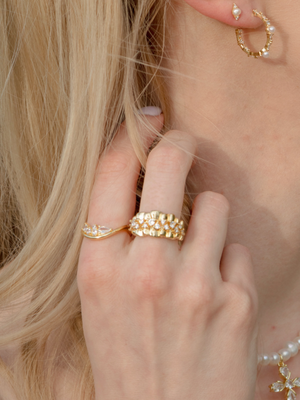 A hand wearing the Kinsey Designs Briony Stone Wave Ring, stacked with other rings, and a gold hoop earring appears near blonde hair and an ear.