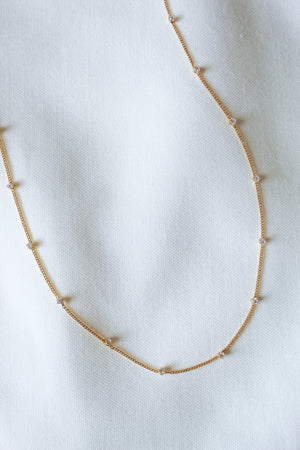 The Bailey Necklace by Kinsey Designs Archives features a delicate 16" gold chain with small pearls, shown on a white textured fabric background.
