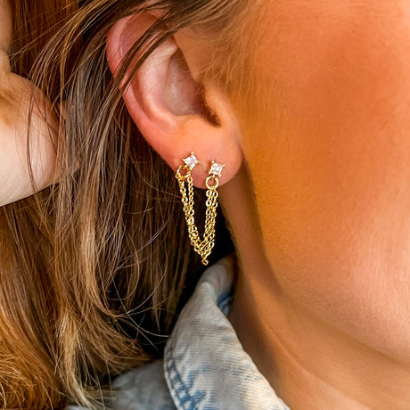 The Becca Double Post by Kinsey Designs Archives features gold-plated chain earrings with two studs, 2" long from post to post, shown on a woman's ear with a partially visible denim collar.