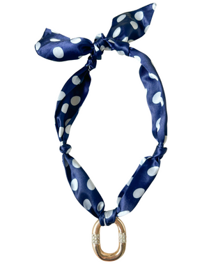 The Bellagio Scarf Necklace by Kinsey Designs features navy blue polka dots and a gold oval pendant with rhinestone accents. This adjustable piece adds timeless charm to any outfit.