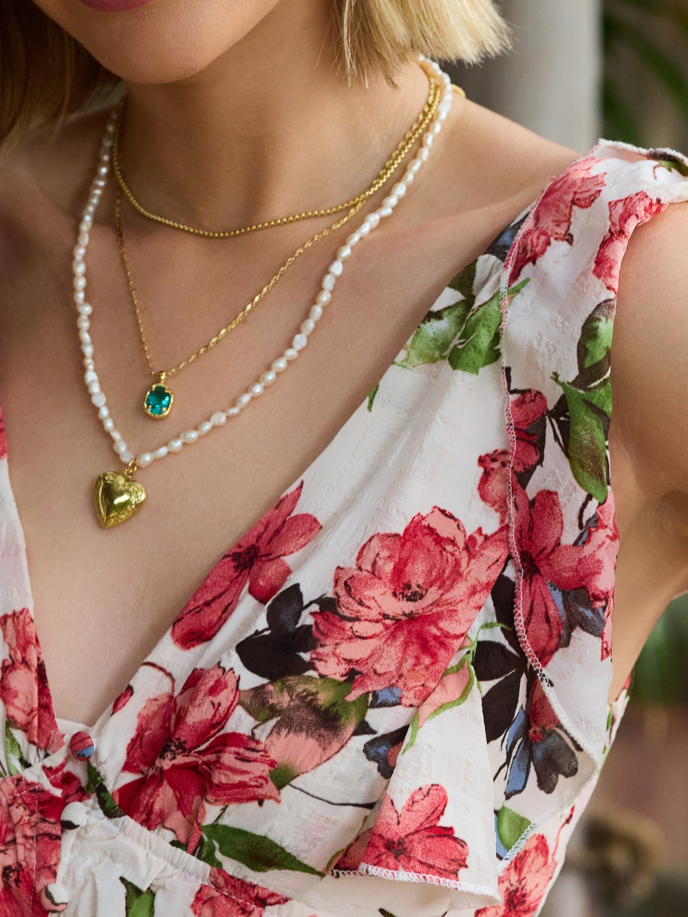 A woman wears layered necklaces, including a gold heart locket and the Kinsey Designs Giselle Pearl Necklace, strung with luminous freshwater pearls, paired with a floral dress.