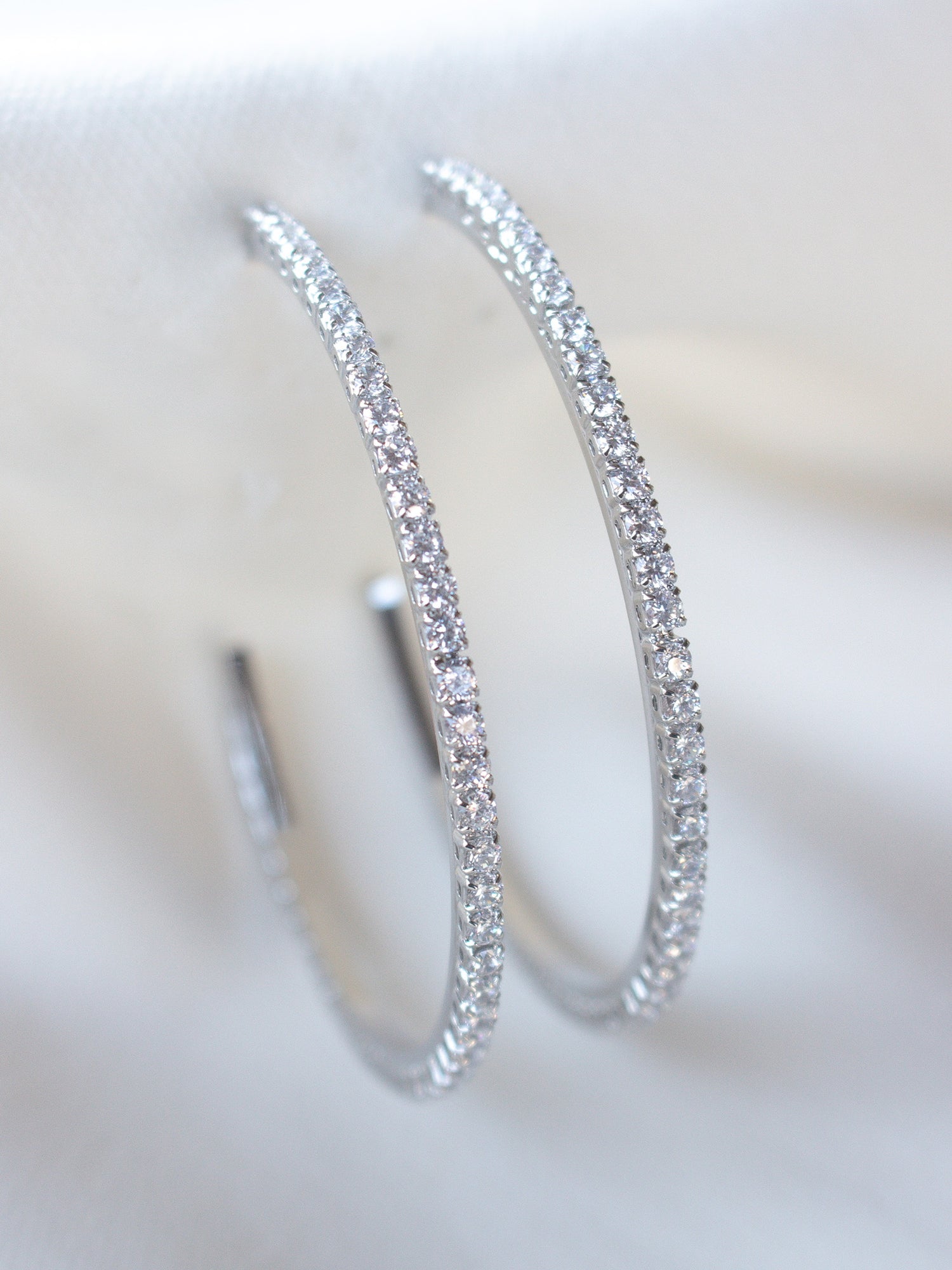 Close-up of Kinsey Designs’ Blaze Hoop Silver earrings, crafted from rhodium-plated brass and adorned with sparkling stones, displayed on white fabric.