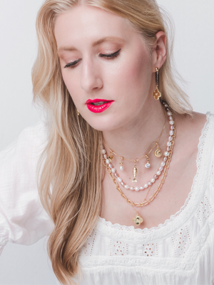A woman with long blonde hair wears a white top, gold earrings, and the Kinsey Designs Bohdi Layering Necklace—a modern chain-and-pearl piece that updates classic necklace layering.