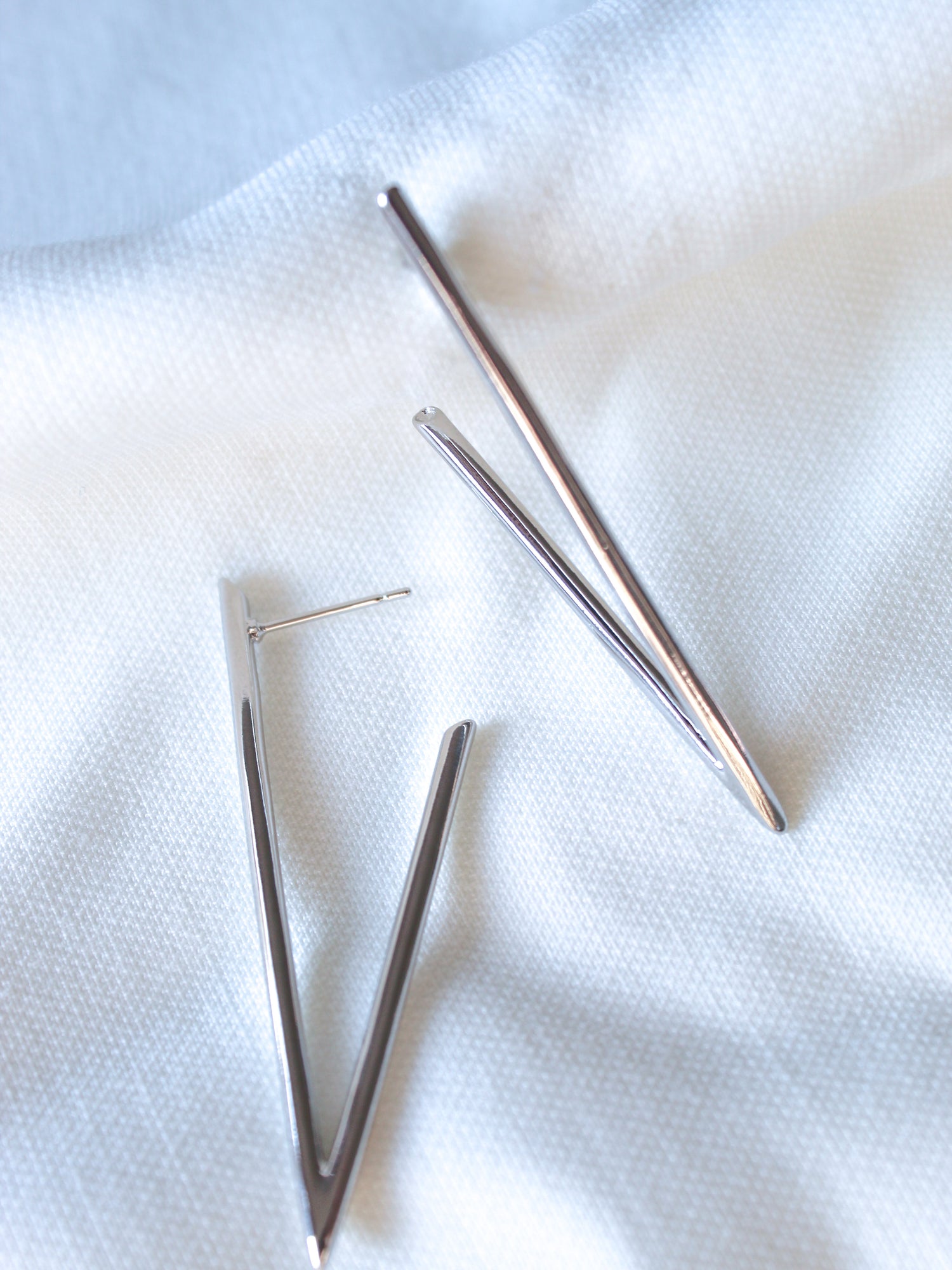 The Bowen Earrings by Kinsey Designs are silver, V-shaped, rhodium-plated earrings shown on a white textured fabric background.