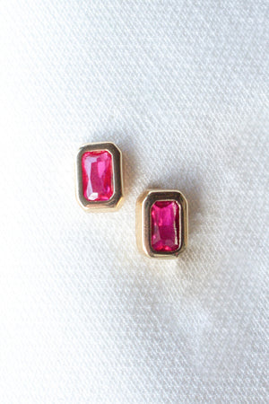 The Brooks Stud earrings by Kinsey Designs Archives, rectangular rubies set in gold, are displayed on white textured fabric and paired elegantly with a sparkling cubic zirconia ring.
