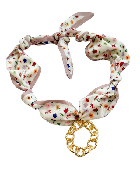 The Calabria Scarf Necklace by Kinsey Designs features a floral fabric with an adjustable design, knotted details, and a large gold chain-link pendant at the center.