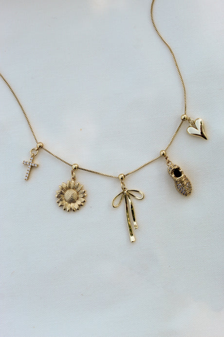 Kinsey Designs' Fancy Bow Grip Charm gold bar necklace features cross, sunflower, textured oval, and heart charms, shown on a white background.