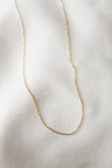 A delicate, thin gold filled chain featuring the NEW! Kinsey Charm Bar Necklace Dainty Cable Chain is elegantly displayed on a white textured fabric background.